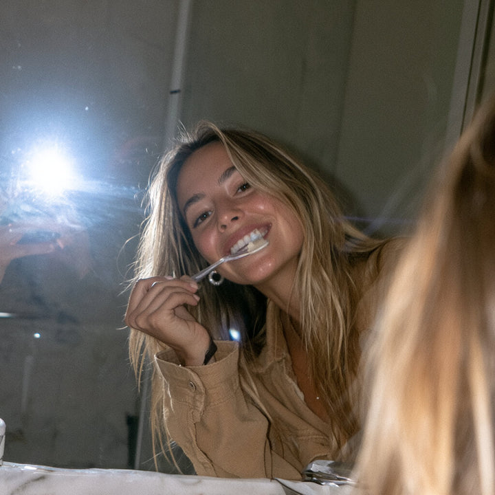 Blonde woman smiling while brushing her teeth in a bathroom mirror selfie